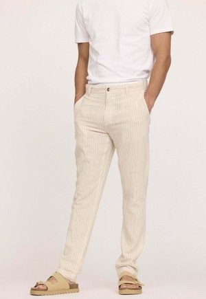 Pantalon rayé beige à coupe slim, avec poches latérales, fermeture par bouton et tissu texturé. Porté avec un t-shirt blanc uni et des sandales.