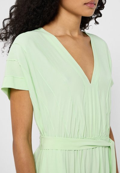Ελαφριά πράσινη φόρεμα με V λαιμόκοψη και κοντά μανίκια, featuring κάθετες πτυχές και στενή μέση με ταιριαστό ζώνη. Λείο υλικό.