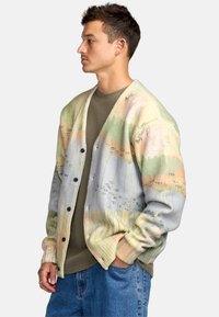 Cardigan van zachte stof met een pastel abstract ontwerp, voorzien van een knoopsluiting aan de voorkant en een relaxte pasvorm. Gecombineerd met een gebreid groen shirt.