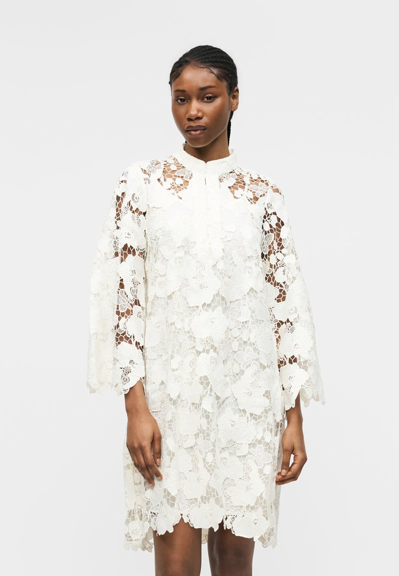 Γυναίκα που φοράει λευκό λουλουδάτο δαντελωτό φόρεμα με μακριά μανίκια, στέκεται μπροστά σε απλό φωτεινό φόντο, κοιτάζοντας μπροστά με ουδέτερη έκφραση.