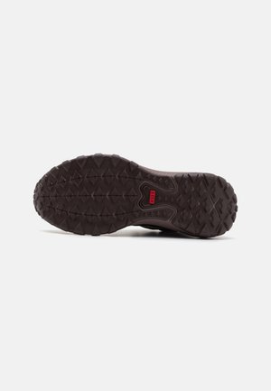 Sole di scarpa sportiva marrone con gomma testurizzata per una buona aderenza. Presenta un motivo pronunciato con lugs di trazione e un accento in rosso nel logo.