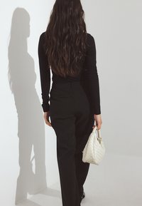 Melna garām piedurknēm blūze un melni bikses, ar baltu austu somu, kurai ir izliekts rokturis, uz gaiša fona.