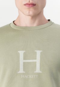 Camiseta de algodón verde oliva con un gran logo blanco de la letra "H" y la palabra "Hackett" debajo en una fuente limpia y sencilla. Cuello redondo.