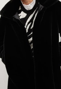 Czarny płaszcz z futra syntetycznego z szerokim kołnierzem, zapięciem na jeden guzik i miękką fakturą. Noszony na czarno-białym swetrze w paski z falbaniastym dekoltem.