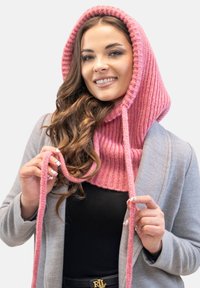 Scialle a cappuccio rosa lavorato a maglia con texture a coste e lacci, indossato sopra un cappotto grigio. Il tessuto sembra morbido e accogliente.