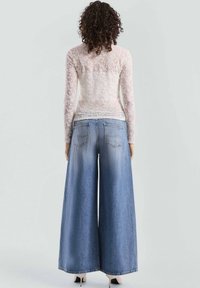 Weißer Spitzenoberteil mit langen Ärmeln, kombiniert mit weitem blauen Denim-Jeans. Das Oberteil hat ein florales Muster; die Jeans haben Gesäßtaschen und ausgefranste Säume.