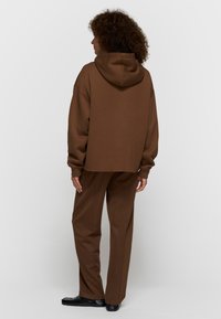 Sweat à capuche marron oversize avec capuche, assorti à un pantalon marron large. Texture lisse et design minimaliste.