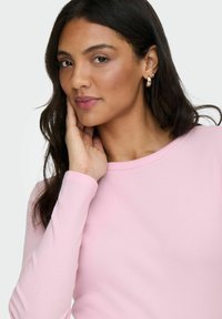 Lichtroze lange mouwen geribbelde top met een ronde halslijn, met een zachte textuur. Het model heeft lang haar en draagt oorringen.