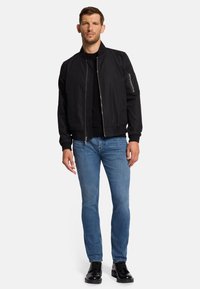 Schwarze Bomberjacke mit Reißverschluss vorne, gerippten Bündchen und einer Reißverschlusstasche am Ärmel, getragen über einem schwarzen Shirt und blauen Jeans.