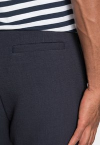 Ciemnogranatowe spodnie o gładkiej fakturze, z kieszenią listwową z tyłu i subtelnym pionowym wzorem. Paski koszuli częściowo widoczne powyżej.