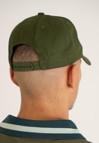 Căciulă verde cu un design structurat, având un sistem de prindere tip snapback, material texturat și un buton circular în vârf. Pare purtată, fiind așezată pe un fundal deschis.