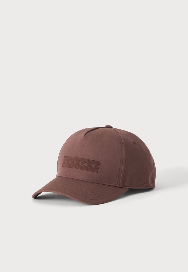 Casquette de baseball Oakley marron avec une visière incurvée et un patch logo cousu à l'avant, présentée sur un fond clair uni.