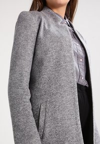 Blazer grigio con tessuto testurizzato, dotato di un colletto alto, tasche laterali e una vestibilità dritta, indossato sopra una camicia con bottoni.