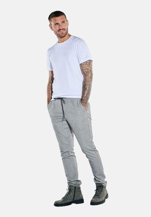 White cotton t-shirt, gray jogger pants with pockets, and dark green lace-up boots. Man stands with a relaxed posture, arms in pockets.