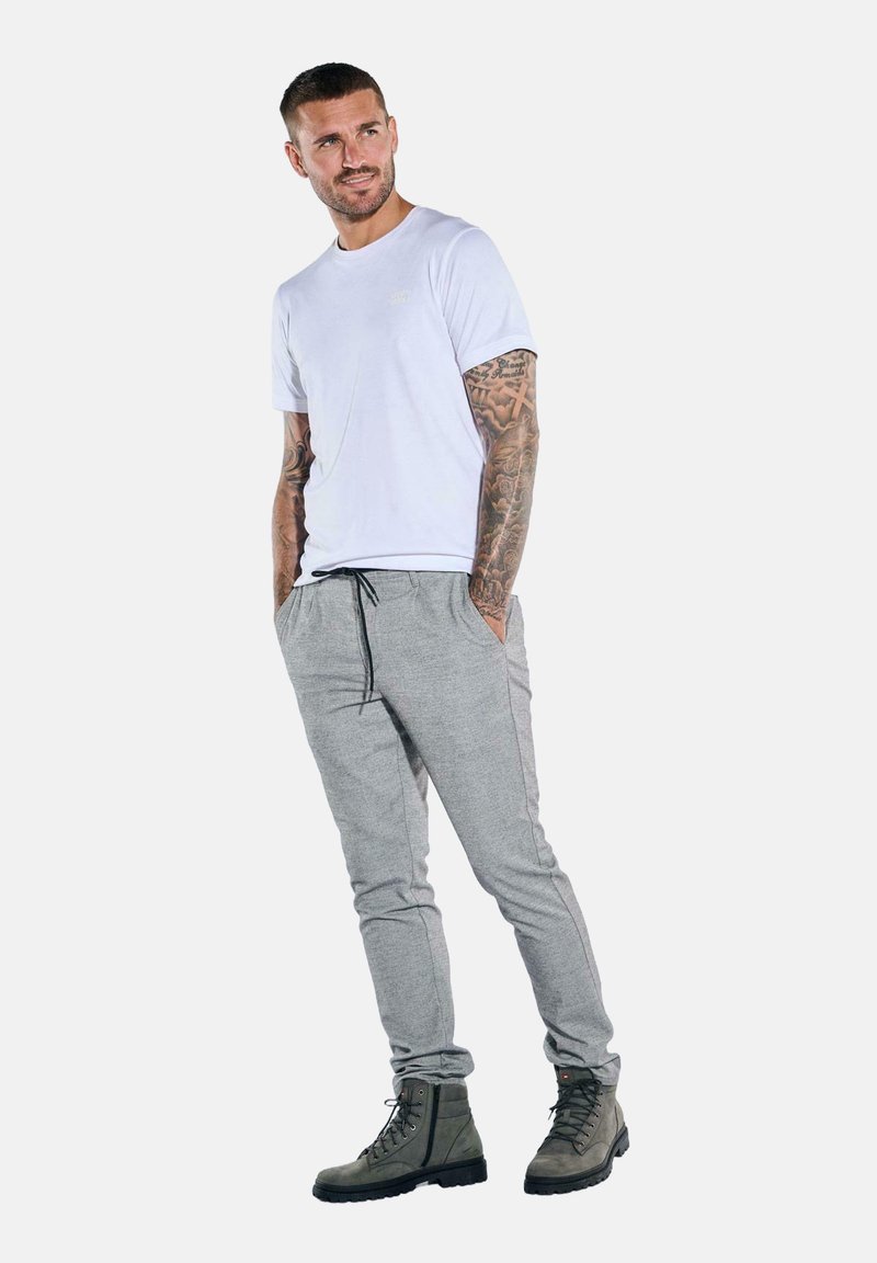 Weißes Baumwoll-T-Shirt, graue Jogginghose mit Taschen und dunkelgrüne Schnürstiefel. Mann steht mit einer entspannten Haltung, die Arme in den Taschen.