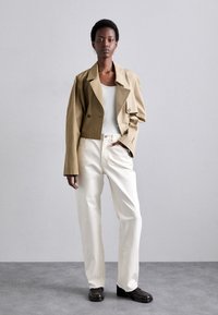 Lyhytnlävinen beige takki, jossa on kaksirivinen muotoilu, yhdistettynä väljään valkoisiin housuihin ja tummiin loafer-kengiin; minimalistinen tyyli.
