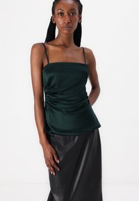 Emerald green satin top with thin straps and gathered detailing at the waist, paired with a black leather skirt, smooth texture.