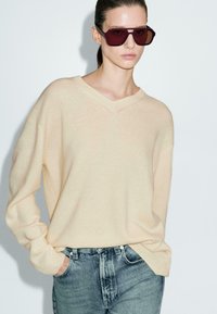 Pull en tricot beige avec un col en V, associé à un jean taille haute bleu clair. Modèle portant des lunettes de soleil oversize bordeaux.