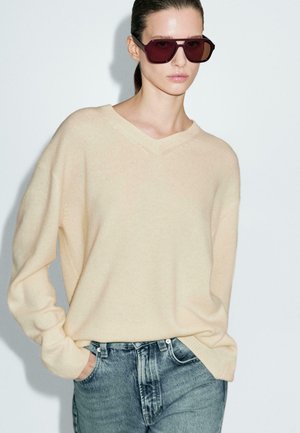 Beige gebreide sweater met een V-hals, gecombineerd met lichtblauwe high-waisted jeans. Model draagt oversized bordeauxrode zonnebril.