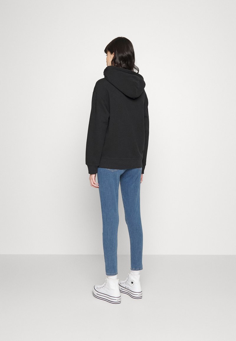 Schwarzer Kapuzenpullover mit Kängurutasche, kombiniert mit hellblauen, figurbetonten Jeans und weißen High-Top-Sneakers. Einfaches und lässiges Design.