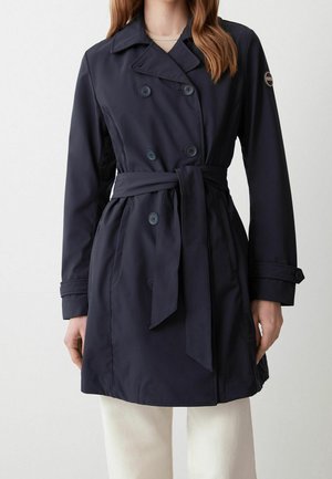 Femme portant un trench-coat bleu marine à double boutonnage avec une ceinture à la taille et des poignets boutonnés, associé à un pantalon de couleur claire.