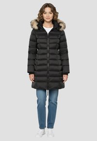 Schwarzer Steppmantel mit abnehmbarem Kapuzenfutter, das mit Fell besetzt ist, ausgestattet mit einem Frontreißverschluss, gerippten Bündchen und einem mittellangen Schnitt. Kombiniert mit blauen Jeans.