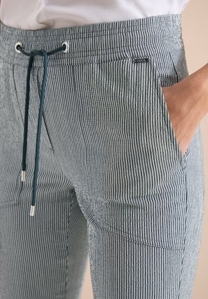 Gros plan d'une personne portant un pantalon rayé vertical gris et blanc avec une taille à cordon et une main dans la poche latérale.