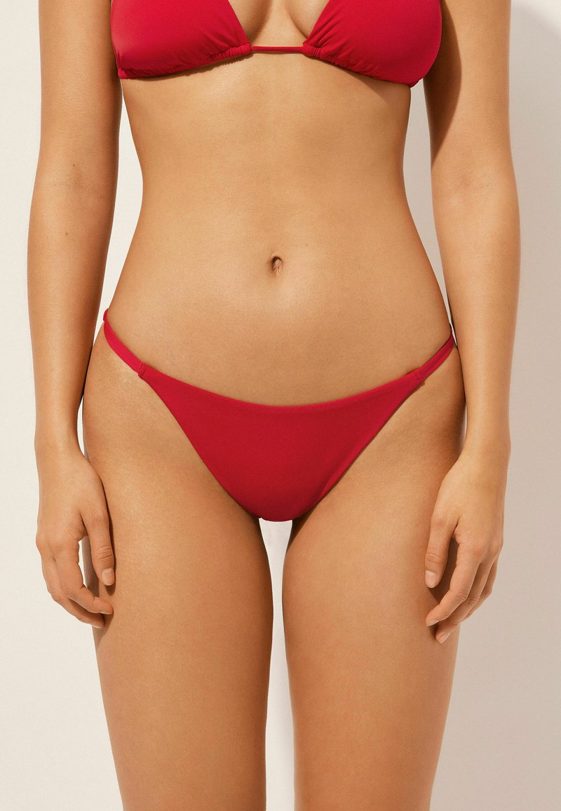 Červený bikiny set s trojúhelníkovým topem a minimálním pokrytím, vyrobený z hladké, elastické tkaniny s tenkými bočními popruhy.