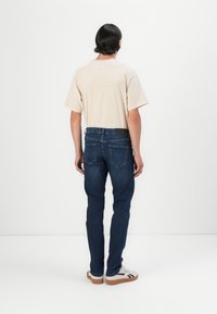 Camiseta beige de manga corta, combinada con jeans de mezclilla azul oscuro de corte recto y con bolsillos traseros. El calzado casual completa el look.