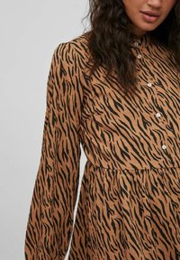 Kobieta ubrana w brązową sukienkę zapinaną na guziki, z długimi rękawami i czarnym wzorem w paski tygrysa, pokazana od ramienia do talii.