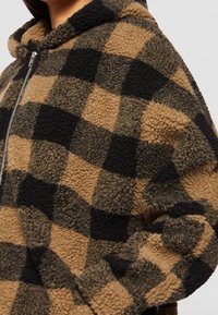 Fleece hoodie i et brunt og sort ternet mønster. Har lynlåslukning og tekstureret stof med et hyggeligt, blødt udseende.