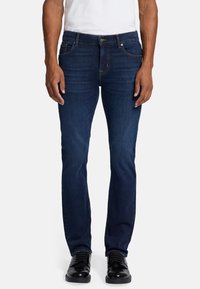 Jeans in denim blu scuro con vestibilità slim, dotati di cinque tasche, cuciture a vista e chiusura con bottone classico. Abbinati a scarpe in pelle nera.