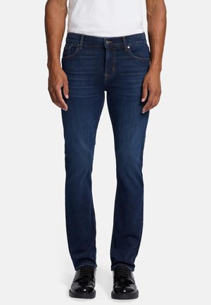 Dunkelblaue Jeans aus Denim mit schmaler Passform, ausgestattet mit fünf Taschen, sichtbaren Nähten und einem klassischen Knopfverschluss. Kombiniert mit schwarzen Lederschuhen.