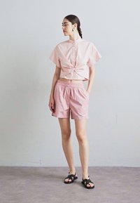Roze cropped shirt met korte mouwen en knoopdetail, gecombineerd met roze gestreepte shorts en zwarte sandalen op een effen achtergrond.