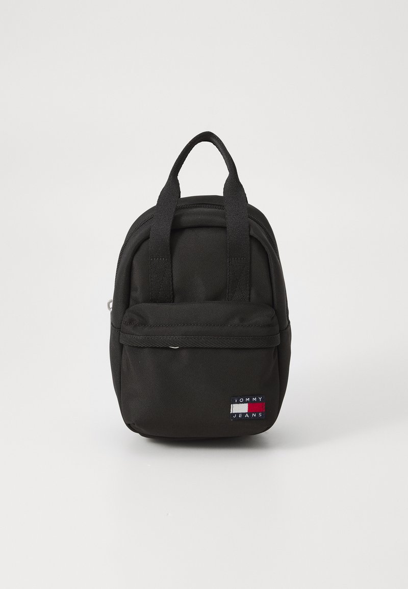 Mini sac à dos en nylon noir avec une poche avant zippée et deux poignées supérieures. Présente une étiquette logo en rouge, blanc et bleu.