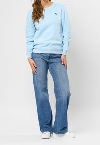 Ljusblå sweatshirt med ribbad mudd och halsringning, ihop med vida blå jeans. Anmärkningslogotyp broderad på bröstet. Vita sneakers.