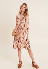 Ein ärmelloses Kleid mit einem orange-, creme- und braunfarbenen Muster, das einen Rüschensaum hat. Kombiniert mit braunen Wedge-Absätzen und einer gewebten Schultertasche.