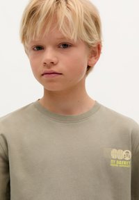 Khakifarbenes Langarmshirt mit Rundhalsausschnitt, das einen gelben Grafikdruck auf der linken Brustseite aufweist und aus glattem Baumwollstoff besteht.