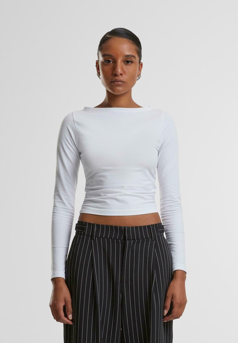 Weißes langärmeliges Crop-Top mit rundem Ausschnitt, getragen mit schwarzen, hoch taillierten Hosen mit feinen Streifen, die eine glatte Textur und eine enganliegende Passform aufweisen.