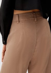 Lichtbruine broeken gemaakt van gladde stof. Bevatten een middelhoge taille, subtiel gestikte tailleband en zijzakken.