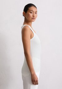 Weißes geripptes Tanktop mit einer taillierten Passform, breiten Schulterträgern und einer glatten Textur, getragen mit weißen Hosen vor einem neutralen Hintergrund.