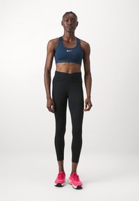 Sötétkék sportmelltartó fehér logóval, fekete, magas derekú leggings-szel és rózsaszín sportcipőkkel párosítva. Simább textúrájú, testhezálló kialakítás.
