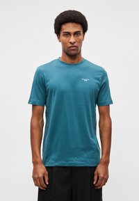 Tricou verde-albăstrui din bumbac, cu guler rotund, mâneci scurte și un mic logo alb pe piept. Textură netedă, croială casual.