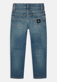 Jeans de mezclilla azul con áreas deslavadas, dos bolsillos traseros, trabillas para cinturón, y un parche negro con el logo de Calvin Klein en el bolsillo derecho.