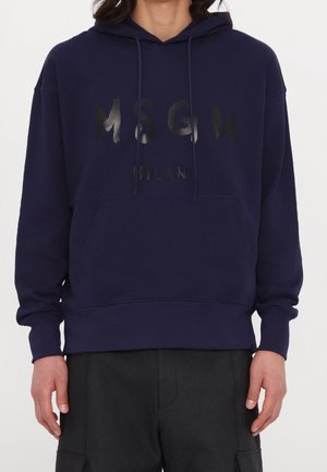 Sweat à capuche bleu marine avec poche kangourou devant et logo MSGM Milan noir brillant, porté par une personne à la carnation moyenne et pantalon noir.