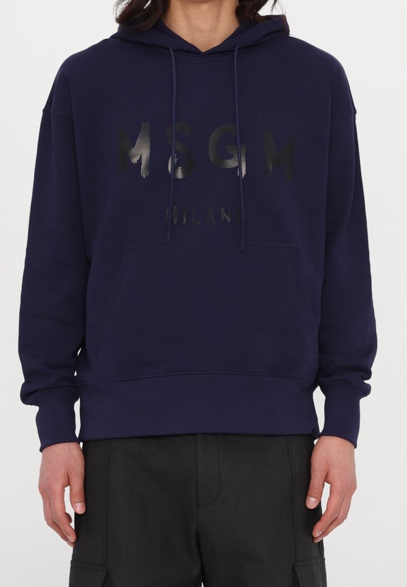 Sweat à capuche bleu marine avec poche kangourou devant et logo MSGM Milan noir brillant, porté par une personne à la carnation moyenne et pantalon noir.