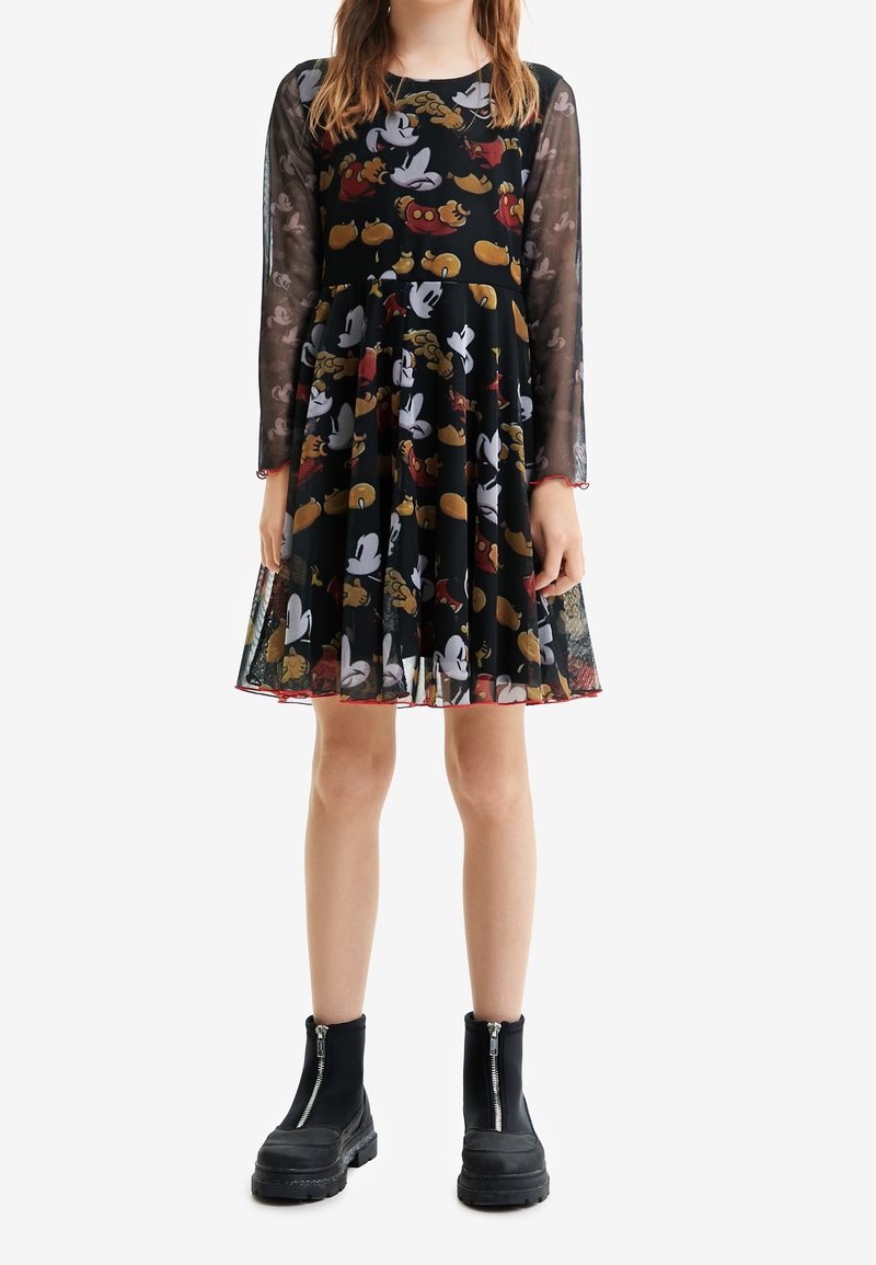 Fille portant une robe noire avec des motifs de gants de dessin animé colorés, manches longues en voile, associée à des bottes noires épaisses à fermeture éclair.