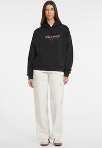 Felpa nera con cappuccio e tasca frontale, testo del logo bianco. Abbinata a pantaloni cargo beige a vestibilità ampia con tasche laterali. Le scarpe da tennis completano il look.