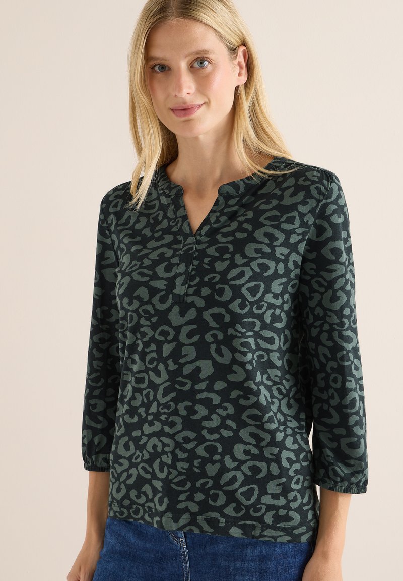 Haut à manches longues de couleur vert foncé avec un motif léopard, doté d'un col à renfoncement et d'un tissu texturé. Associé à un jean bleu.