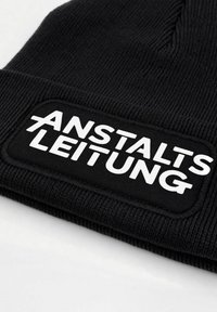 Black knit beanie featuring a rectangular patch with white text "ANSTALTS LEITUNG". Text is in a bold, sans-serif font.